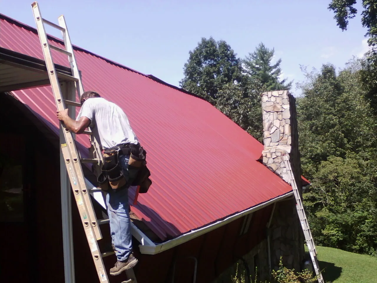 Roofer on a ladder beside a red metal roof and stone chimney — Soffit & Fascia Repair by Roofing Brothers in Only
